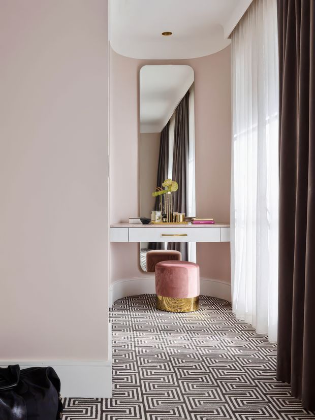 A refined dressing or entry nook with a wall-mounted white console beneath a tall rounded-rectangle mirror. The space has blush-pink walls and a curved ceiling detail, full-height windows dressed with sheer white curtains and dark brown drapes, and a bold geometric black-and-white patterned carpet. A plush pink velvet stool with a gold-toned base sits in front of the console, creating a glamorous, softly layered interior.