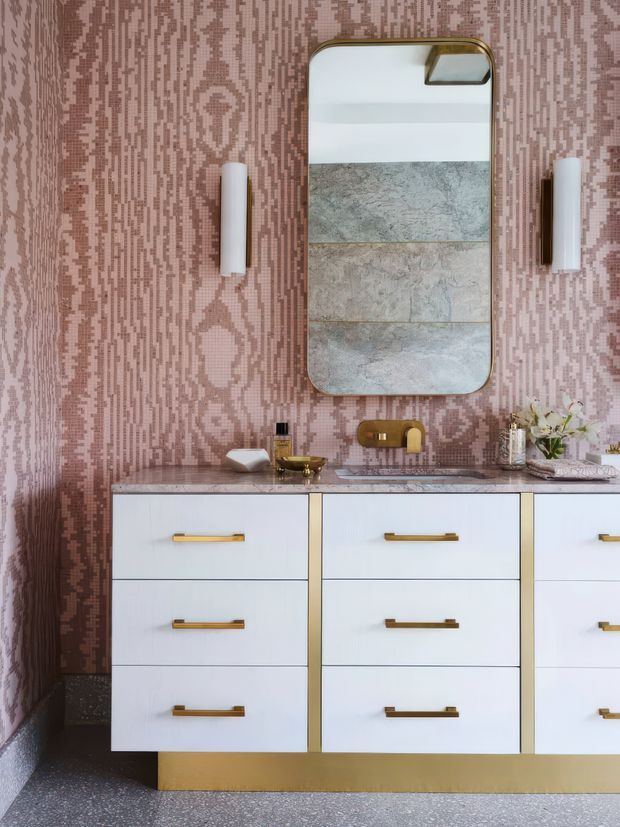 A richly styled bathroom vanity vignette featuring a wide white built-in cabinet with six drawers, brass handles and brass plinth, topped with a mottled stone benchtop and gold-toned wall-mounted tap. The wall is covered in a pink and mauve patterned tile or textured finish, with a rounded rectangular brass-framed mirror and matching vertical wall lights. Decorative toiletries and a small floral arrangement sit on the vanity, creating a glamorous, artful interior.
