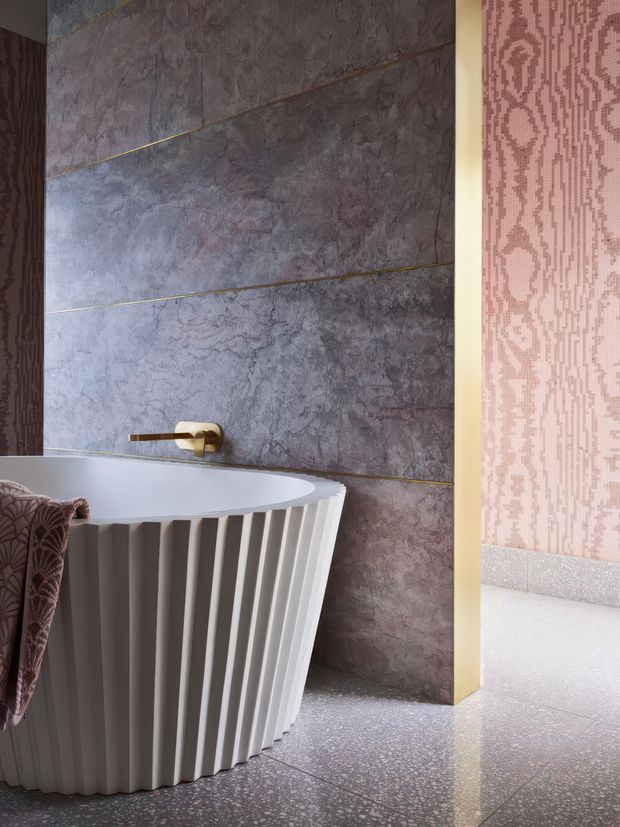 A stylish bathroom featuring a freestanding white bathtub with a vertically fluted exterior, set against large dark grey-purple stone-look wall panels with thin brass-toned joints. A wall-mounted brass tap is positioned above the bath. The floor is a light terrazzo-like surface, and a patterned mauve-pink wall covering is visible through the opening to the right. The space has a refined, dramatic, contemporary character.