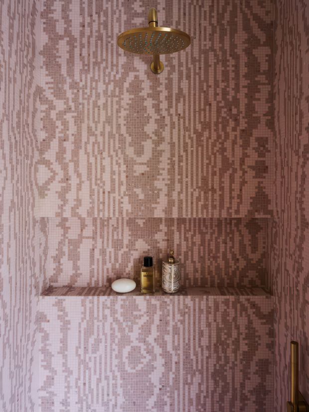 A compact shower enclosure clad entirely in small pink and dusty mauve mosaic tiles arranged in an irregular vertical pixel-like pattern. A round brushed brass showerhead and controls provide a warm metallic accent, while a recessed tiled niche holds soap and toiletry bottles. The space is intimate, highly patterned, and softly lit.