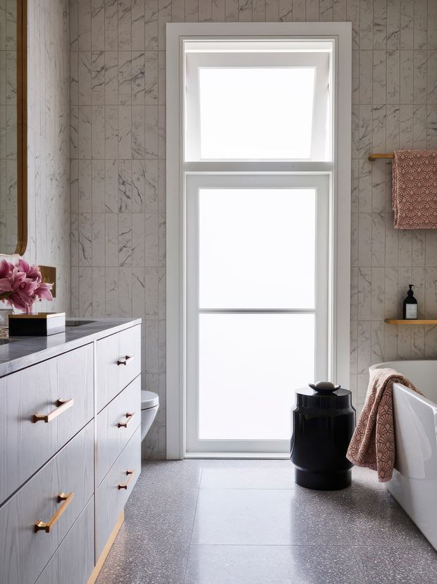 Bright, refined bathroom interior with a large white-framed window, pale marble-look tiled walls, a light wood vanity with brass hardware and dark benchtop, terrazzo-style flooring, and a freestanding white bathtub partially visible at right. Warm patterned towels, timber shelves, and a black vessel add contrast.