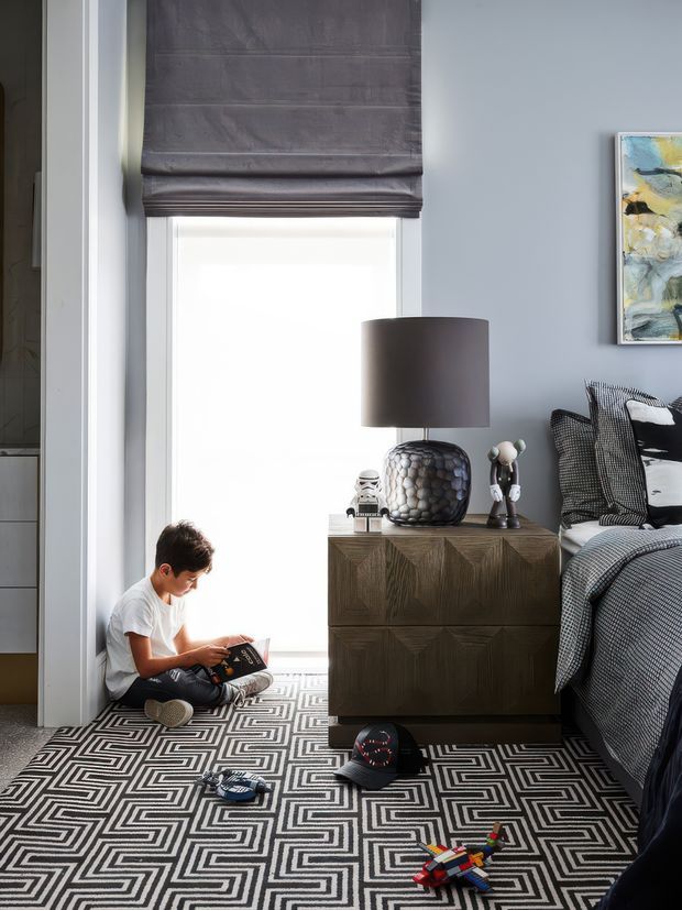 A stylish children's bedroom with a monochromatic geometric patterned carpet, soft grey-blue walls, a tall window with a dark Roman blind, and a dark timber bedside cabinet. The bed is dressed in layered grey and black patterned textiles, with a sculptural lamp and decorative objects on the cabinet. A child sits reading beside the window, while toys and personal items are scattered on the floor.