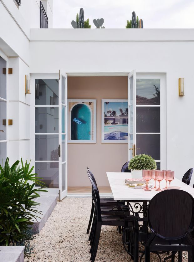 A bright white architectural courtyard viewed from an outdoor dining area. The enclosed courtyard has white rendered walls, tall glazed French doors opening into an interior room, brass wall sconces, framed artwork, gravel paving, and sculptural cactus planting on the roofline. A white dining table with black metal chairs and pink glassware occupies the foreground, while leafy potted plants soften the space.