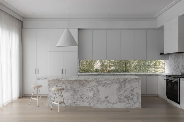 A refined contemporary kitchen with full-height pale cabinetry, a large marble-clad island, integrated appliances, a suspended white pendant light, and a long horizontal window overlooking trees. Light timber flooring, sheer curtains, and restrained detailing create a bright, calm interior.
