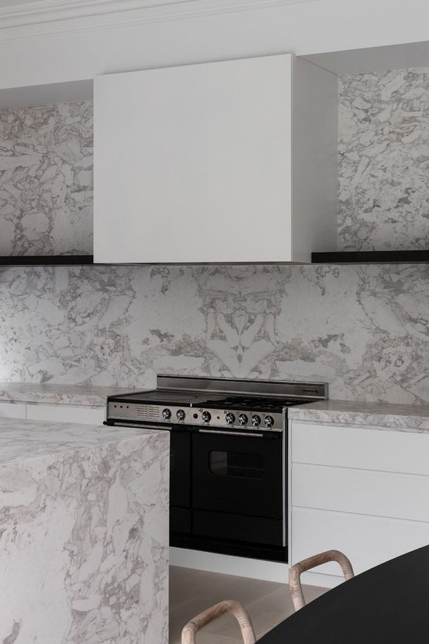 A contemporary kitchen featuring extensive white and grey veined stone surfaces across the splashback, benchtops and island cabinetry surrounds. White flat-panel cabinetry, a large white rangehood, black built-in oven and cooktop, and a dark island benchtop create a high-contrast, refined scheme. Pale timber bar stools are partially visible in the foreground.
