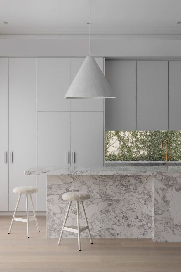 A refined minimalist kitchen with full-height pale grey cabinetry, a large veined stone island and matching benchtop, a slim horizontal window with greenery beyond, and a brushed brass tap. Two light upholstered stools sit beneath a sculptural white pendant light. The space has a soft, monochromatic palette with warm timber flooring and subtle natural texture.
