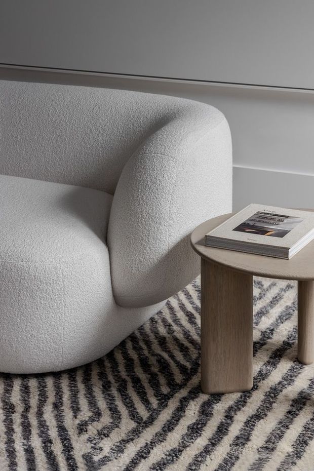Close-up interior vignette featuring a rounded, sculptural off-white upholstered armchair beside a small round light-wood side table with a book. A bold black-and-cream patterned rug covers the floor, with simple light neutral wall panelling in the background. The styling is calm, refined, and contemporary with softly curved forms.