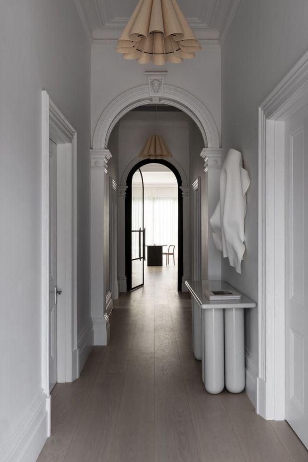 A refined, monochromatic residential hallway with pale grey walls, white decorative mouldings, paneled doors, ornate arched openings, and a light timber floor. Sculptural pendant lights hang along the corridor, while a slim console and abstract wall sculpture add contemporary accents. A bright room is visible at the far end.