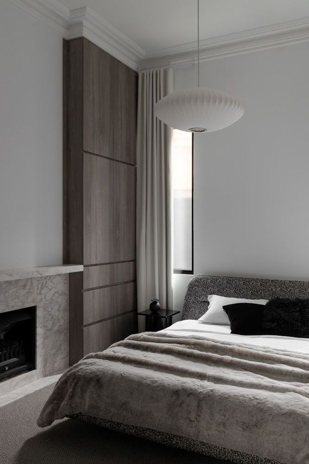 A refined bedroom with a dark timber built-in wardrobe, upholstered bed, layered grey bedding, black and white cushions, full-height curtains, ornate ceiling cornices, and a suspended sculptural pendant. A pale marble fireplace forms a feature at the left, creating a sophisticated monochromatic interior.