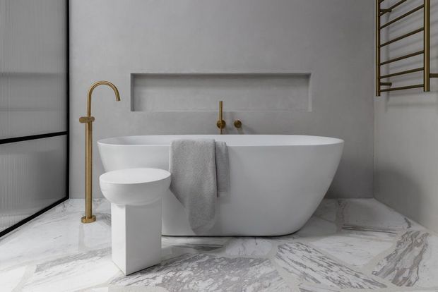A refined contemporary bathroom featuring a freestanding white bathtub, sculptural pedestal basin, brushed brass floor-mounted tapware, wall-mounted brass fixtures, a recessed wall niche, heated towel rail, and dramatic grey-veined marble flooring. The palette is predominantly white and grey with brass accents.