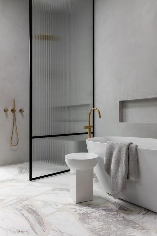 A refined contemporary bathroom featuring a freestanding white bathtub, sculptural pedestal basin, brushed gold wall-mounted shower fittings and tapware, a black-framed fluted glass shower screen, pale textured walls, and dramatic white-and-grey marble flooring. A recessed wall niche provides additional storage.