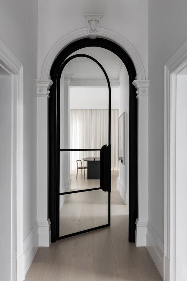 A refined white architectural hallway with ornate classical architrave mouldings and a dramatic black steel-framed glazed arched door. Pale timber flooring leads into a bright adjoining room with sheer curtains, a small round table, and a chair. The image combines traditional detailing with a contemporary black-framed pivot or hinged door.
