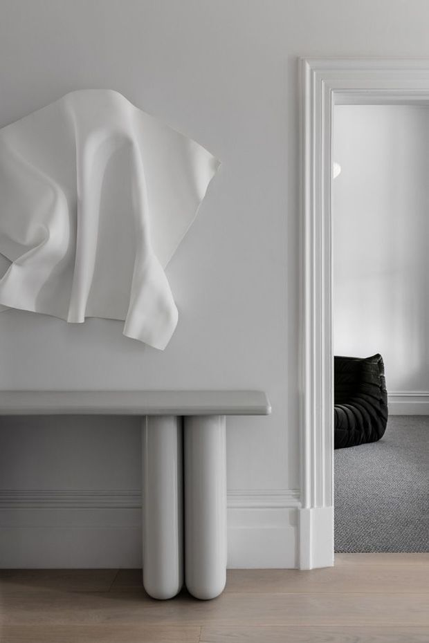 A refined monochromatic interior vignette featuring a sculptural white console table with rounded cylindrical legs against a pale grey wall with decorative wall mouldings. An abstract draped white wall sculpture hangs above the console. Through the wide, heavily trimmed doorway, a softly furnished adjacent room is visible with a black curved lounge chair and grey carpet. Pale timber flooring and crisp white architectural trims complete the calm, minimalist composition.