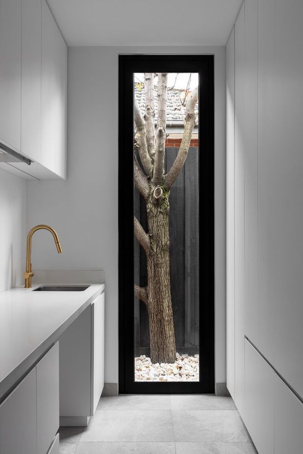 A narrow, minimalist utility or laundry room with white built-in cabinetry on both sides, a white benchtop and inset stainless-steel sink, and a brass gooseneck tap. A tall black-framed glazed door or window provides a dramatic view of a tree growing in a small white-stone courtyard. The room has pale tiled flooring, white walls and ceiling, and a clean monochromatic palette with brass detailing.
