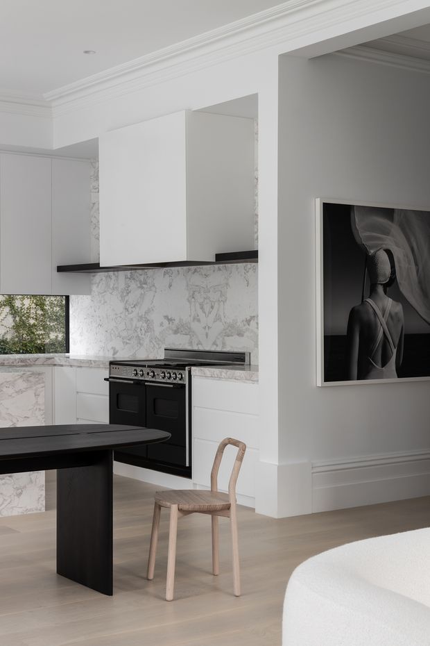 A refined open-plan kitchen and dining area in a predominantly white interior. The kitchen features white cabinetry, dramatic white-and-grey veined stone splashbacks and benchtops, a black freestanding range cooker, and dark island table. Pale timber flooring, a light timber chair, decorative wall moulding, ceiling cornices, and a large monochrome artwork create a sophisticated, restrained setting.