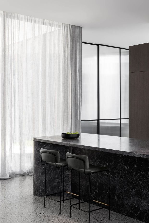 A refined contemporary kitchen with a dark veined stone island and matching waterfall side, slim black-framed internal glazing, sheer full-height curtains, and two black bar stools with brass footrests. The space has a monochromatic palette, polished stone surfaces, and terrazzo flooring.