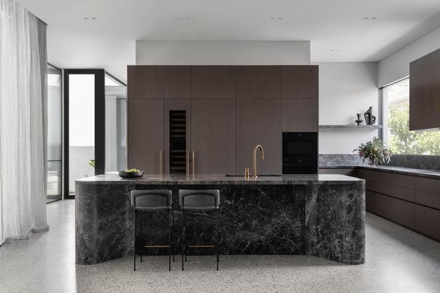 Contemporary kitchen with a dramatic dark stone island, integrated dark timber cabinetry, built-in appliances, brass tapware, and two bar stools. Light terrazzo-style flooring, sheer curtains, and generous glazing create a refined, modern interior with strong contrast between the pale shell and dark joinery.