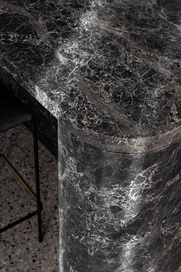 Close-up detail of a dark veined natural stone countertop or rounded island edge, with a polished black and grey marble surface. A terrazzo floor and part of a black metal-framed stool are visible in the background.