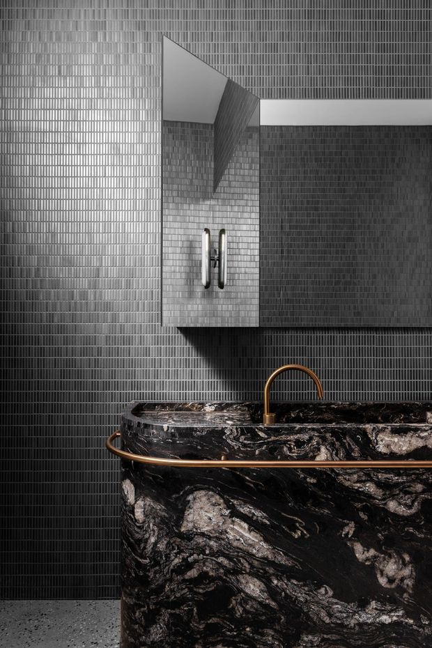 A dramatic, close-up view of a sophisticated bathroom vanity. Dark vertically ridged wall tiles surround a floating grey-toned cabinet with integrated handles and a mirrored or reflective upper surface. A sculptural black stone basin with bold white veining dominates the foreground, paired with a curved brass tap and matching brass rail detail. The space has a moody, luxurious, highly textural contemporary feel.