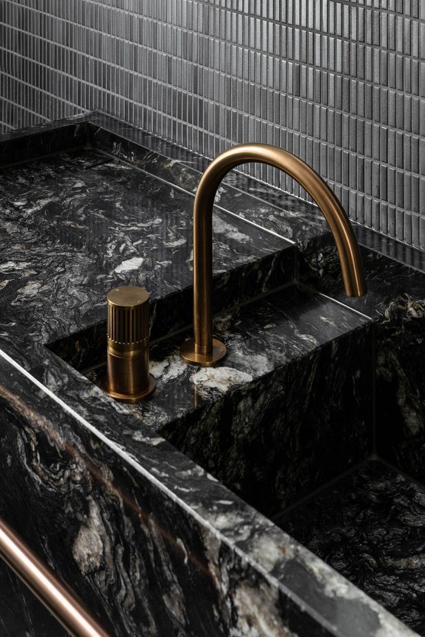 Close-up of a dramatic dark stone bathroom basin with a sculptural bronze gooseneck tap and matching cylindrical mixer. The basin and surrounding surfaces are clad in richly veined black marble or natural stone, while the backsplash features small vertically ridged dark grey tiles. The composition is luxurious, moody, and highly material-focused.