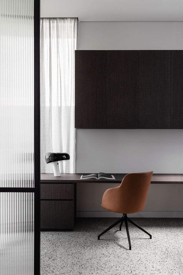 A refined home office with a long built-in dark timber desk and matching wall-mounted cabinetry. A brown upholstered swivel chair sits at the desk beside an open book and a small lamp. Sheer curtains filter daylight through a window, while a vertically ribbed glass partition frames the left side. The room has a pale grey wall, dark joinery, and a light terrazzo-style floor.