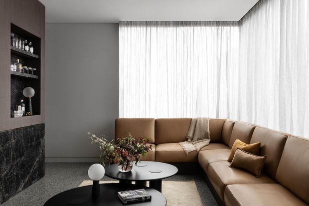 Refined contemporary living room with a large tan L-shaped sectional sofa, layered round coffee tables, and floor-to-ceiling sheer curtains wrapping around a bright corner window. A dark stone feature wall with integrated shelving and a sculptural globe lamp contrasts with the soft neutral furnishings and grey carpet.