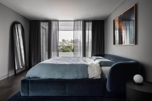 A contemporary bedroom with a large upholstered dark blue bed, matching curved lounge chair, dark timber flooring, and charcoal-grey walls. Full-height sheer and blackout curtains frame a bright window, while an arched full-length mirror and framed artwork add visual interest. The room has a refined, moody atmosphere with clean lines and soft textures.