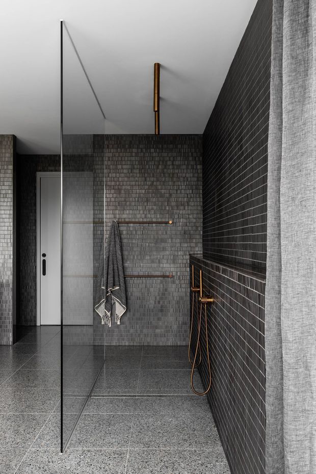 A contemporary walk-in shower bathroom with dark grey small-format tiled walls, matching large-format floor tiles, a frameless glass shower screen, and copper-toned shower fixtures and towel rails. A grey towel hangs on the rear wall, with a white internal door visible at the back. The space has a restrained, monochromatic palette with warm metallic accents.