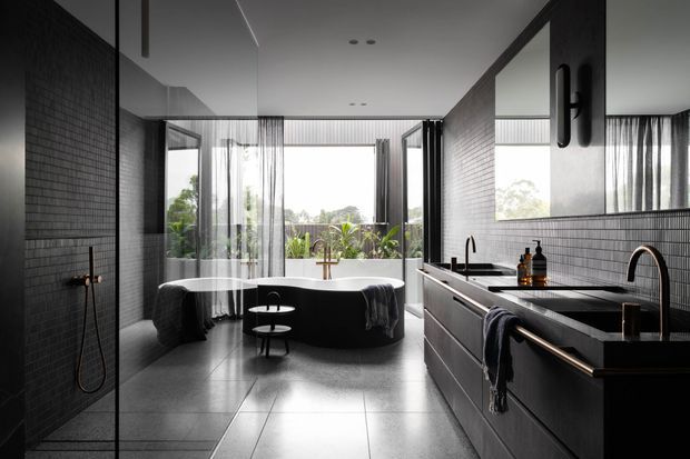 A sophisticated contemporary bathroom with a freestanding oval bathtub positioned in front of floor-to-ceiling glazing, a walk-in shower behind a glass partition, and a long dark vanity with integrated basins and wall-mounted fixtures. Dark tiled walls, large-format grey floor tiles, sheer curtains, and black metal details create a moody, refined atmosphere. Garden greenery is visible outside the windows.