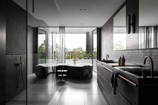 A contemporary, generously sized bathroom with a freestanding oval bathtub positioned in front of floor-to-ceiling glazing. The room features dark small-format tiled walls, large grey floor tiles, a long dark vanity with integrated basins and wall-mounted mirrors, a glass-enclosed shower, and black tapware. Sheer curtains and greenery outside soften the otherwise monochromatic, refined interior.