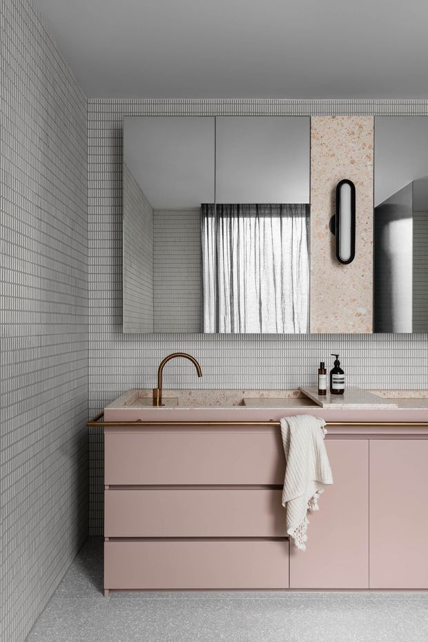 A refined contemporary bathroom vanity featuring full-height small-format grey grid wall tiles, a dusty blush-pink floating vanity with slim brass detailing, a pale terrazzo or stone benchtop with integrated basin, brushed brass tapware, and a large mirrored cabinet. A sculptural black wall light and sheer curtain add contrast and softness.
