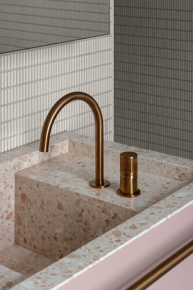 Close-up of a refined bathroom vanity featuring a sculptural bronze gooseneck faucet and matching cylindrical tap set into a thick pink-and-cream terrazzo countertop and integrated basin. The backdrop is composed of pale vertically ribbed tiles, with a soft pink vanity front and bronze trim creating a warm, decorative finish.