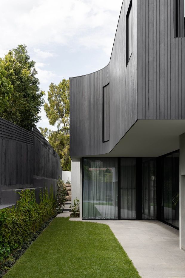 A contemporary residential exterior viewed from a narrow landscaped courtyard. The house features dark vertical timber cladding, a sculptural curved upper facade with a cantilevered overhang, tall black-framed glazed doors, and a restrained lawn-and-paving landscape bordered by a dark privacy fence.