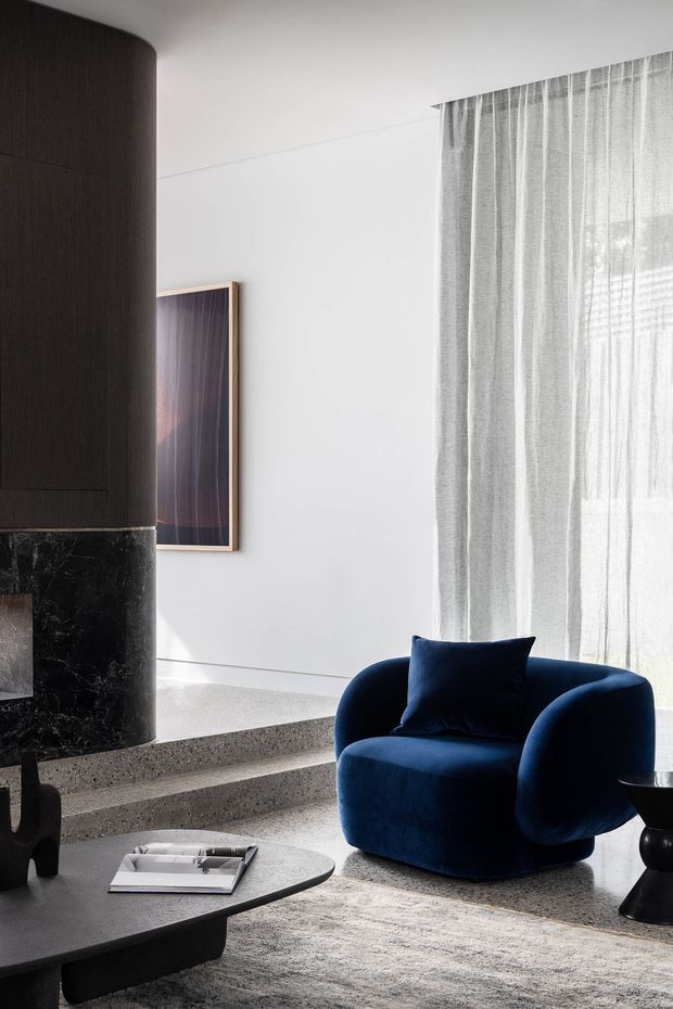 A refined contemporary living room with a sculptural dark curved fireplace surround, black marble base, terrazzo steps and flooring, sheer floor-to-ceiling curtains, and a deep navy boucle armchair. A low dark coffee table, neutral rug, and framed artwork complete the restrained, luxurious interior.