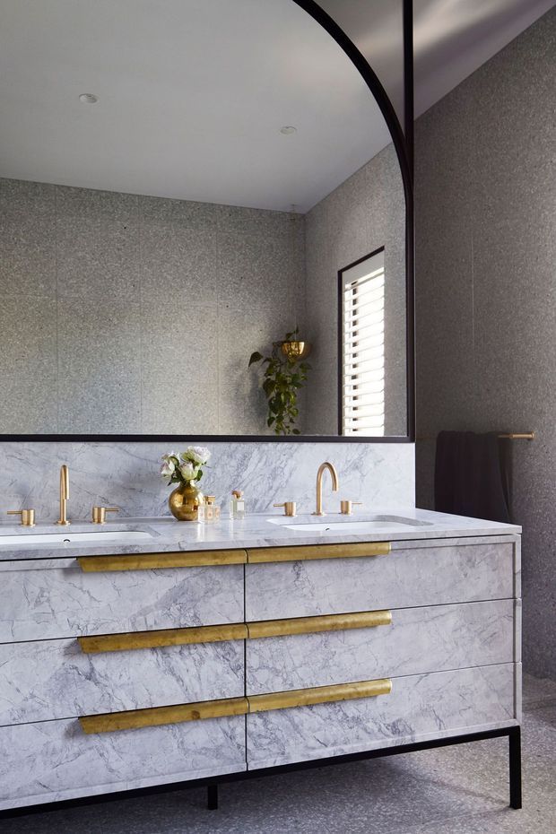 A refined bathroom vanity with double basins, pale veined marble-effect cabinetry and benchtop, and prominent brushed brass hardware and tapware. The room features dark grey textured wall and floor finishes, a large black-framed mirror, white ceiling, and a shuttered window. A small floral arrangement and toiletries sit on the vanity.