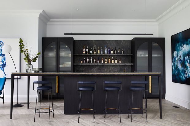 A sophisticated home bar and entertaining area with a long dark timber island and four slim bar stools. The room features black built-in cabinetry with glass-front display cupboards, open shelves stocked with bottles, a dark stone-look backsplash, brass accents, white walls with decorative cornices, herringbone timber flooring, contemporary artwork and sculptural lighting.