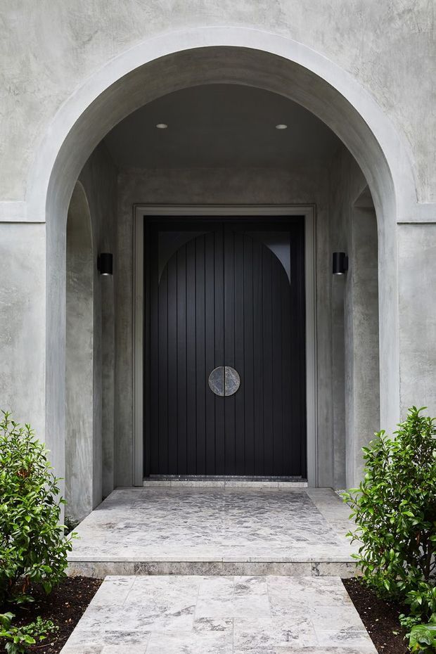 A dramatic residential entrance viewed from outside, featuring a tall black vertically paneled door with a circular metal handle set within a deep rounded concrete or rendered archway. The facade and entry surround have a rough grey finish, with recessed ceiling lights and slim black wall sconces. A pale stone-paved path leads to the doorway, framed by clipped green shrubs.
