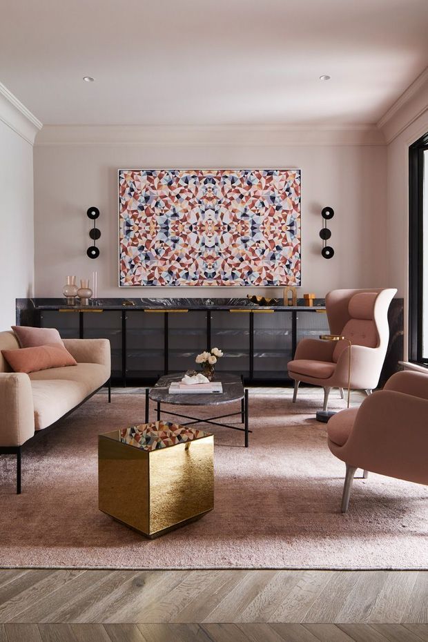 Elegant, highly styled living room with soft blush-beige walls, ornate ceiling cornices, and a large geometric abstract artwork above a long dark console. The room features sculptural mid-century-inspired lounge chairs and sofa seating, a black metal-framed coffee table, a reflective gold cube side table, layered neutral rugs, and pale timber flooring. Tall black-framed glazing is visible at right, while recessed ceiling lights and decorative wall sconces provide restrained illumination.
