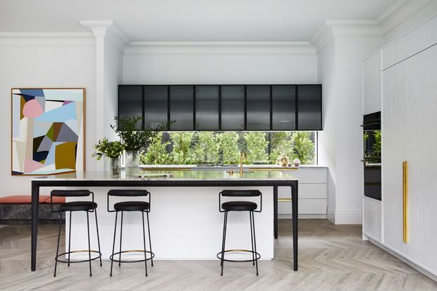 Bright contemporary kitchen with a large white island, black-framed bar stools, dark roller blinds, extensive white cabinetry, marble-look benchtops, and a full-height window overlooking greenery. The room features decorative wall panelling, classical cornice and column details, herringbone timber flooring, and a colourful abstract artwork.