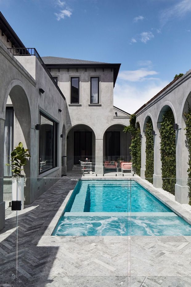 A refined internal courtyard with a narrow rectangular swimming pool, surrounded by rendered masonry walls and repeated arched openings. The courtyard includes dark stone or marble-look paving in a herringbone pattern, a glass pool safety barrier, climbing greenery, black-framed windows and doors, and a two-storey rendered building with a shallow hipped roof. Bright sunlight and blue sky create a crisp, elegant atmosphere.