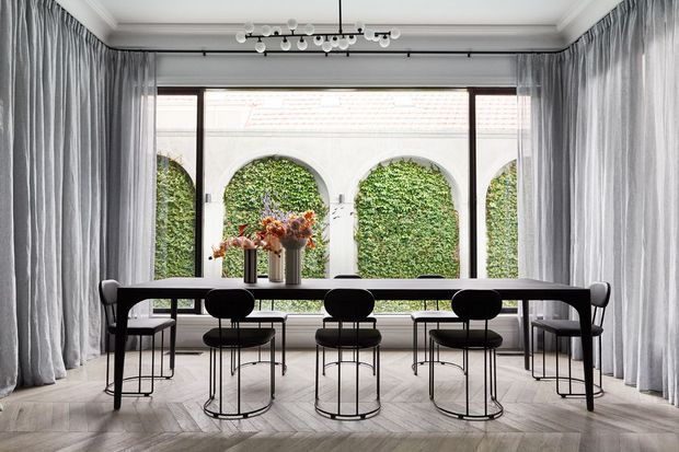 A refined dining room with a long dark dining table and matching black chairs arranged before a large glazed opening. Full-height grey curtains frame the windows, which overlook a lush courtyard with arched openings and greenery. The room features pale herringbone timber flooring, white decorative ceiling mouldings, and a sculptural modern pendant light.