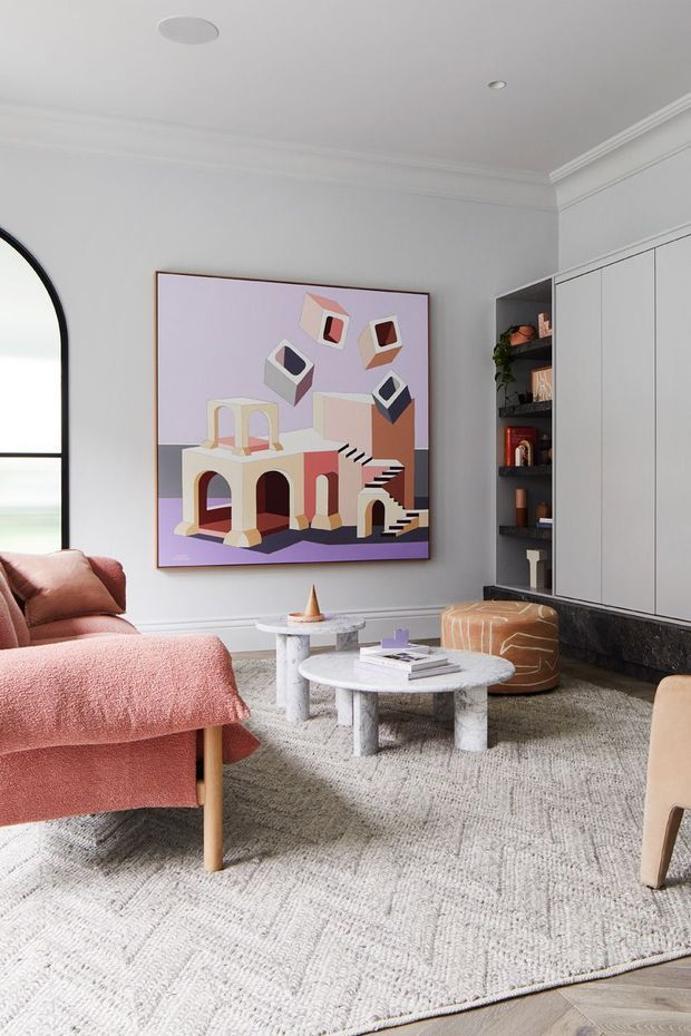 A refined living room with soft white walls, ornate ceiling cornices, built-in white cabinetry and dark stone detailing. The room features blush-pink upholstered lounge chairs, a textured pale rug, sculptural round coffee tables, a leather ottoman, and a large colourful abstract artwork. An arched black-framed window and open shelving add architectural and decorative interest.