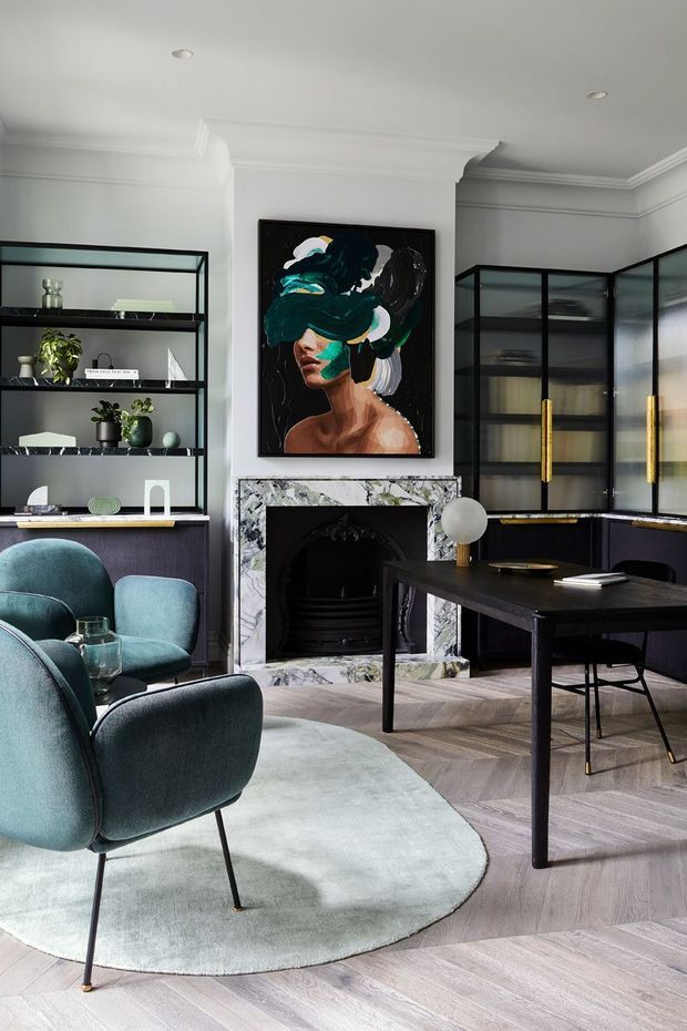 A refined open-plan living and dining room with a black marble fireplace, dark built-in display shelving and glazed cabinetry, a black dining table, teal upholstered lounge chairs, and a large abstract artwork. The room features pale grey walls with decorative cornices, herringbone timber flooring, and a soft light-grey rug. The overall design is elegant, moody, and contemporary with classic detailing.