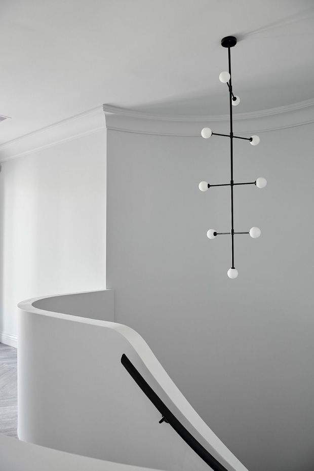 Minimalist interior view of a curved stairway and landing, featuring a smooth white plaster balustrade with a black handrail, crisp decorative ceiling cornices, and a sculptural black linear pendant with spherical white lights.