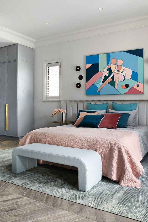 A contemporary bedroom with a soft grey upholstered bedhead, layered blush pink, teal and burgundy bedding, and a curved upholstered bench at the foot of the bed. The room includes grey built-in cabinetry with a brass-toned vertical handle, light timber flooring, pale walls with decorative cornices, a shuttered window, and a colourful geometric artwork above the bed.