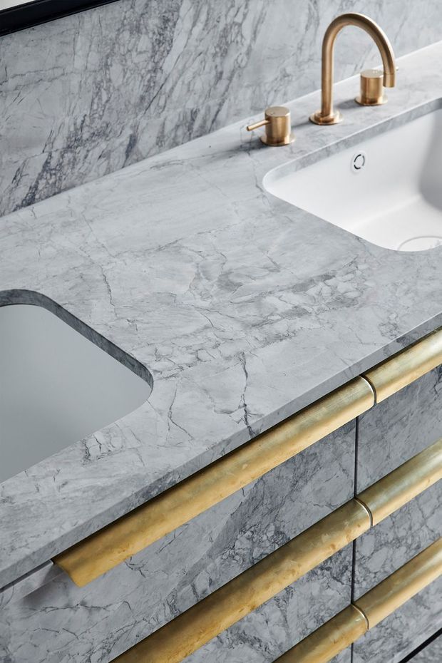 Close-up view of a refined bathroom vanity featuring a grey veined marble countertop and matching marble wall and cabinetry surfaces. The integrated white basin is paired with a curved brushed brass/gold tap and cylindrical handles. Rounded brass drawer pulls add a sculptural detail to the built-in vanity.