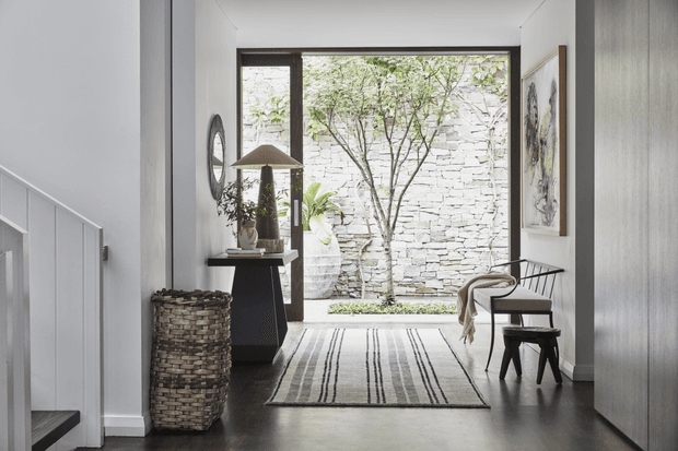 A refined, minimalist entry hall with dark timber flooring, white walls, and a large glazed opening to a small stone courtyard. A staircase with white balustrade is visible on the left, while a console table, woven basket, artwork, chair, stool, and striped rug furnish the space.