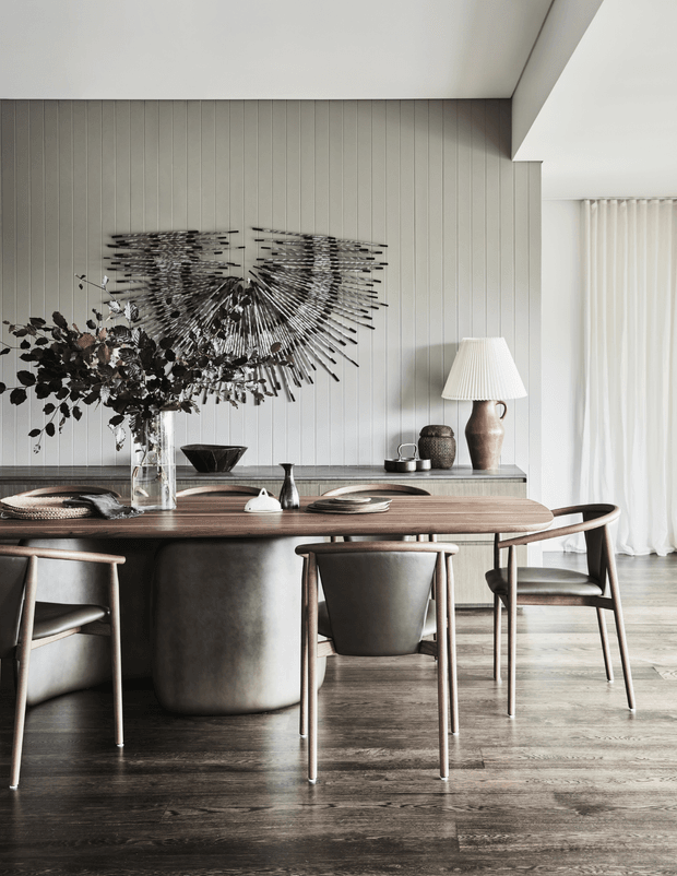 A refined dining area with a long rounded-edge timber dining table, sculptural dark pedestal base, and four upholstered timber-framed dining chairs. The room features dark timber flooring, vertically panelled grey wall cladding, a low sideboard, sculptural wall art, and a softly lit table lamp beside sheer white curtains. The styling is warm, contemporary, and understated.