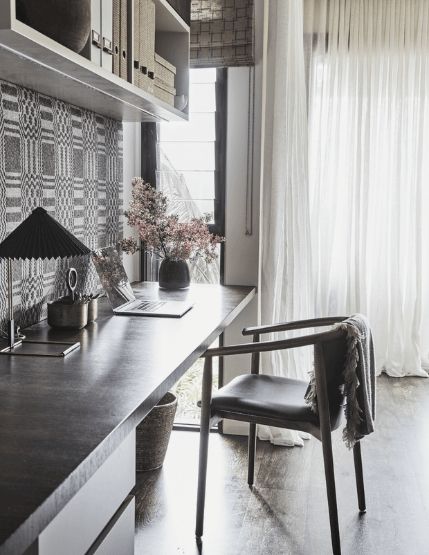 A refined home office nook with a long dark built-in desk beneath open shelving. The space features patterned monochrome wall covering, woven storage baskets, a black table lamp, decorative ceramics, a laptop, and a dark wood-and-leather chair beside full-height sheer curtains and a tall window. The overall look is warm, minimalist, and sophisticated with strong natural light.