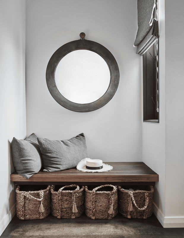 A compact, refined entryway nook with a built-in dark timber bench, four woven storage baskets underneath, grey cushions, and a large round aged-metal framed mirror. A narrow window with a textured Roman blind provides natural light, while the neutral palette and rustic materials create a calm, inviting feel.
